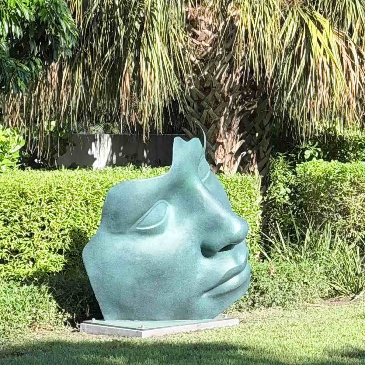 Large bronze abstract face sculpture with green patina displayed among hedges in a landscaped garden