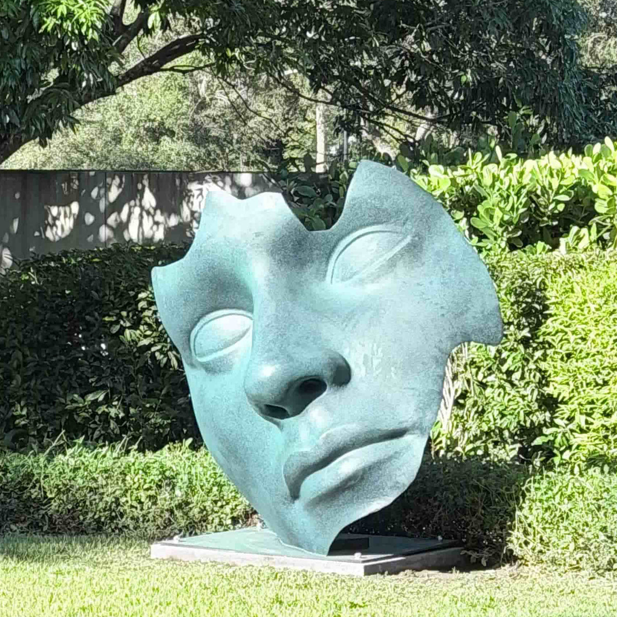 Outdoor bronze abstract face sculpture with green patina installed in a garden, side view