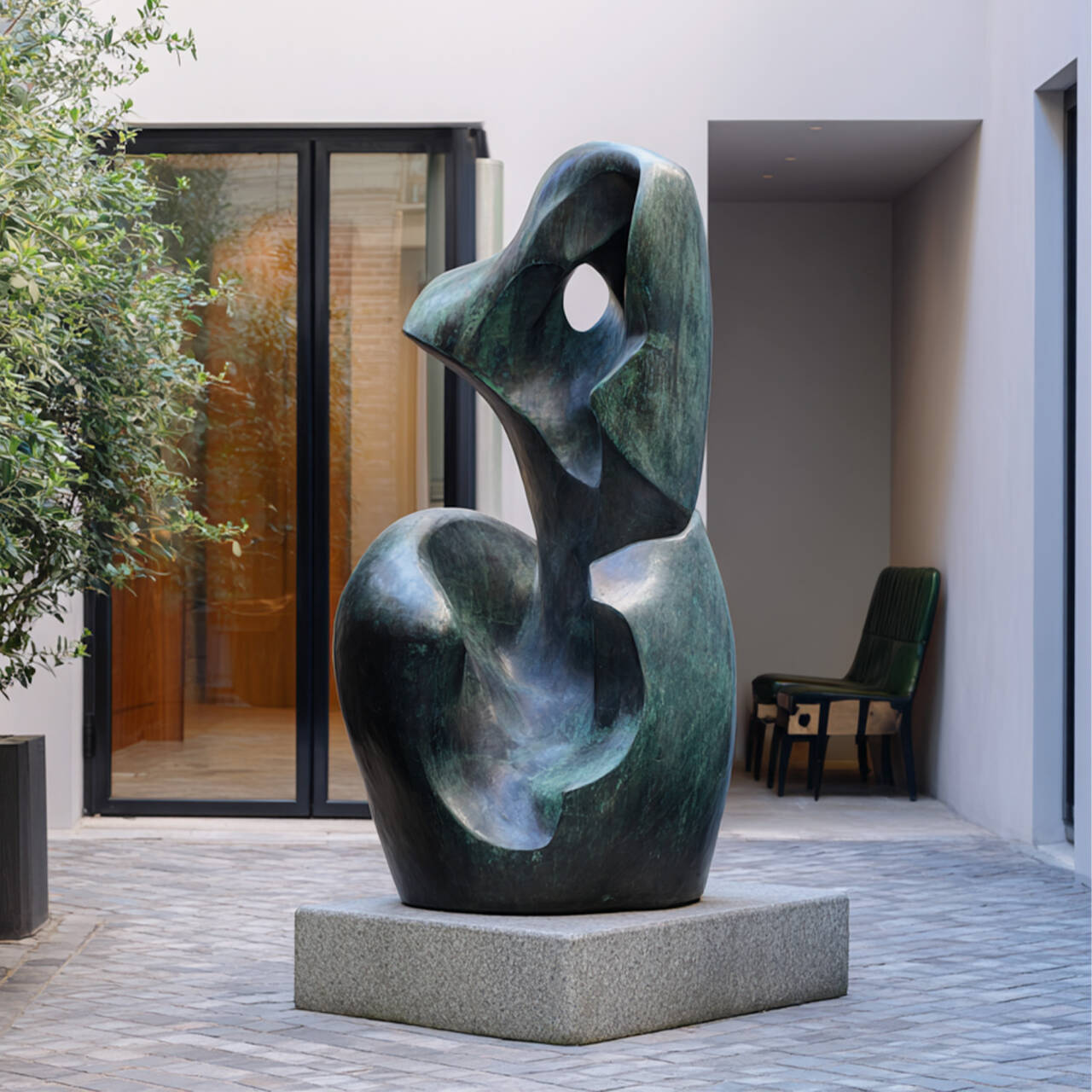 Large verdigris bronze abstract outdoor garden sculpture on a stone base, installed in a modern courtyard with glass doors and stone paving