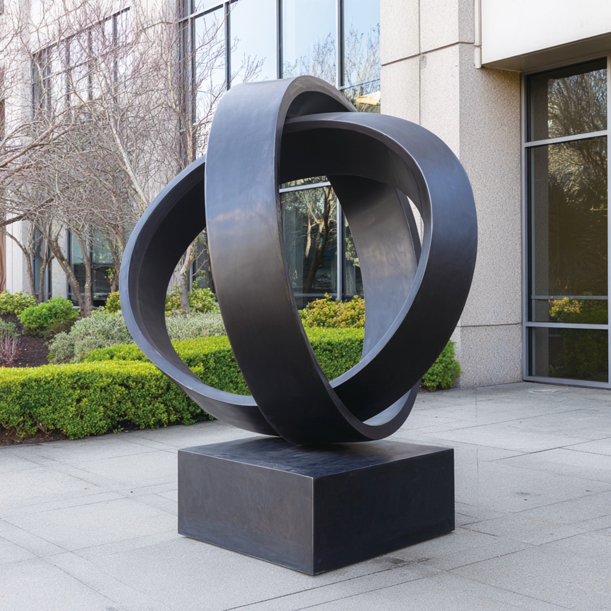 Bronze modern abstract sculpture with intertwined rings, displayed in an outdoor setting near a sleek modern building, ideal for enhancing contemporary lawns or public spaces