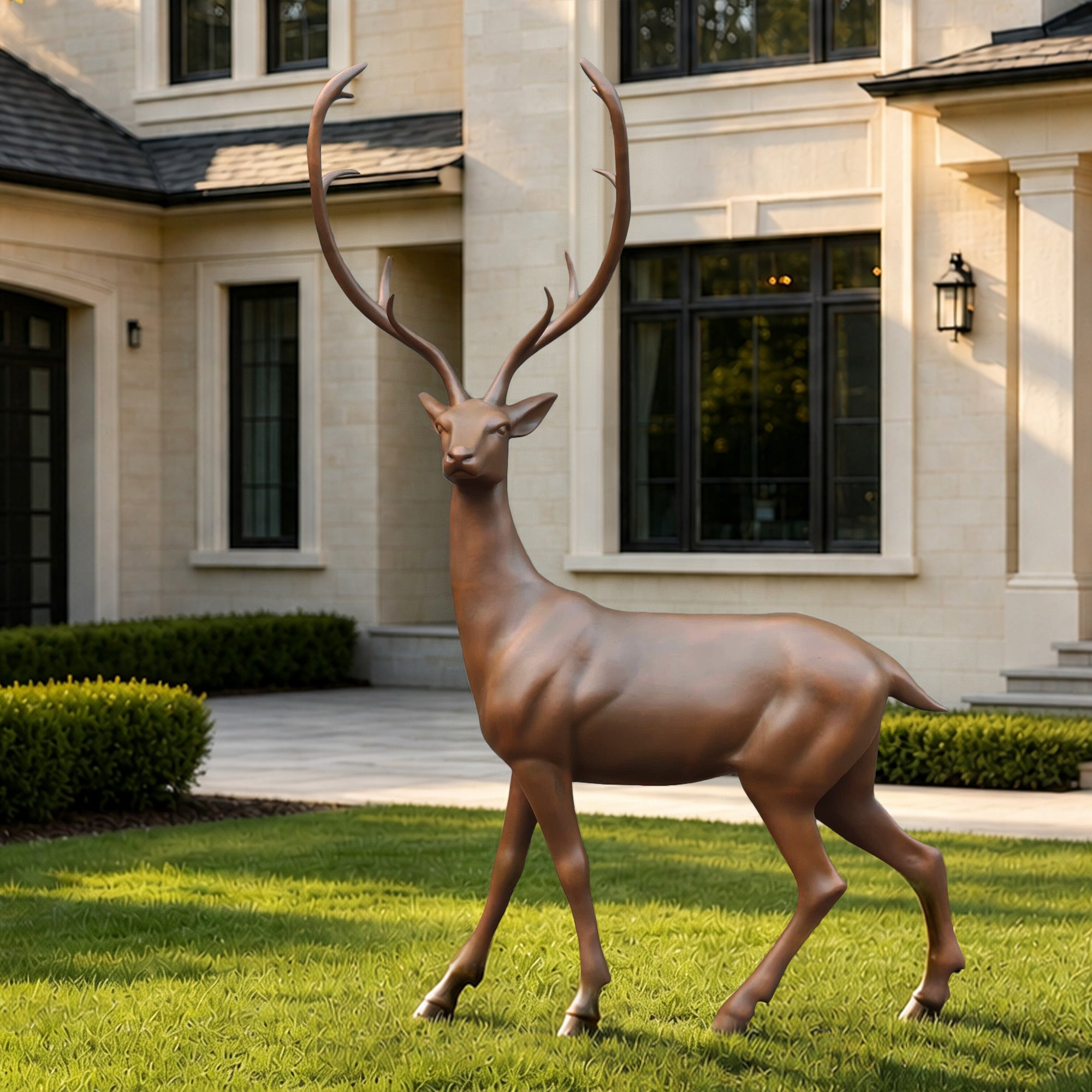 Life-size bronze stag sculpture displayed on a manicured lawn outside a luxury residence, featuring a graceful walking pose, tall branching antlers, and a warm bronze finish.