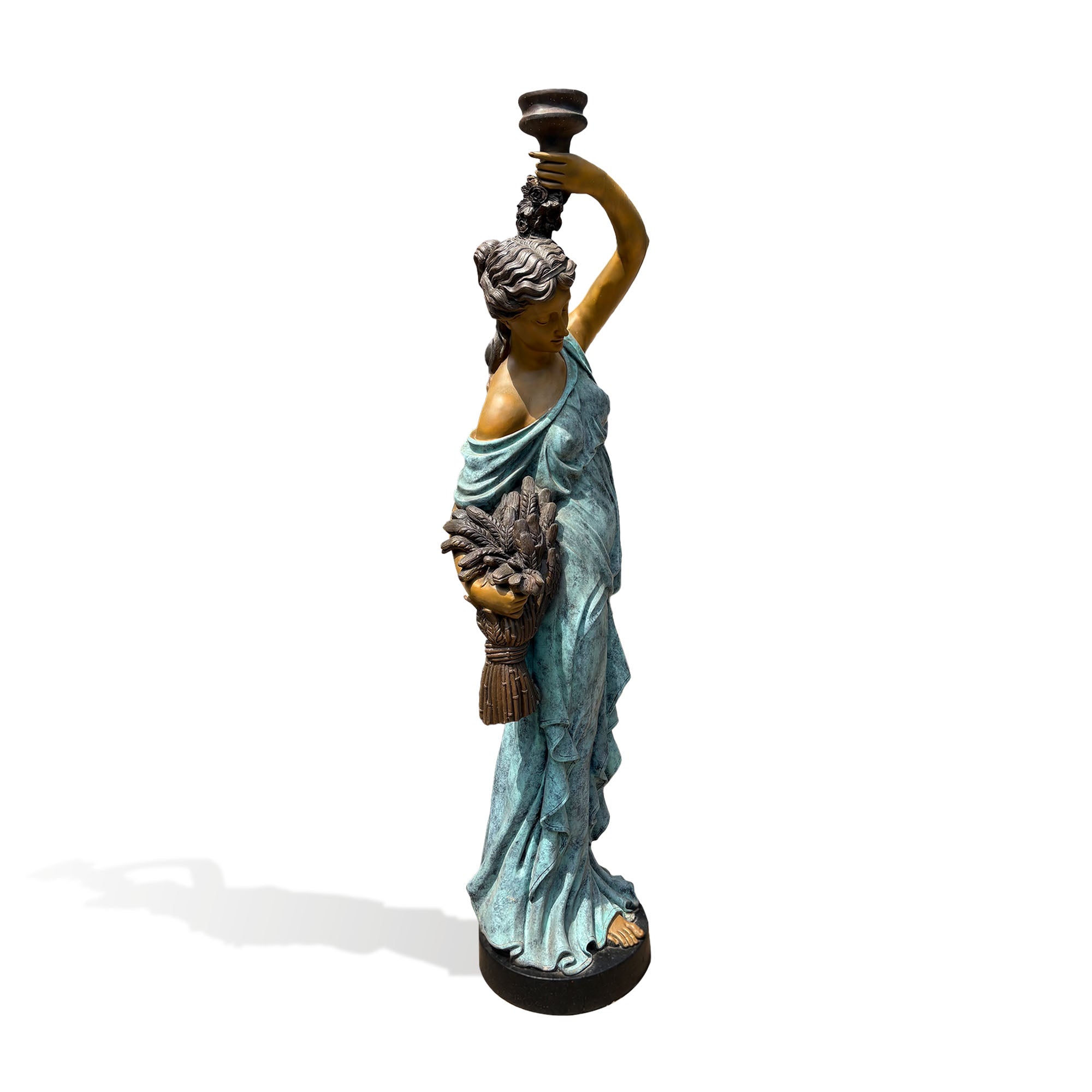Three-quarter view of a classical bronze woman sculpture with verdigris patina finish, raised ceremonial vessel, detailed drapery, harvest sheaf, and circular base.