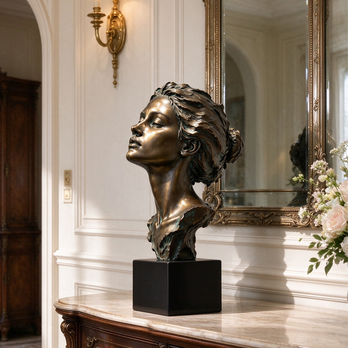 Bronze-finished female bust sculpture on a black rectangular base, displayed on a marble console table in an elegant interior with a mirror, wall sconce, and flowers.
