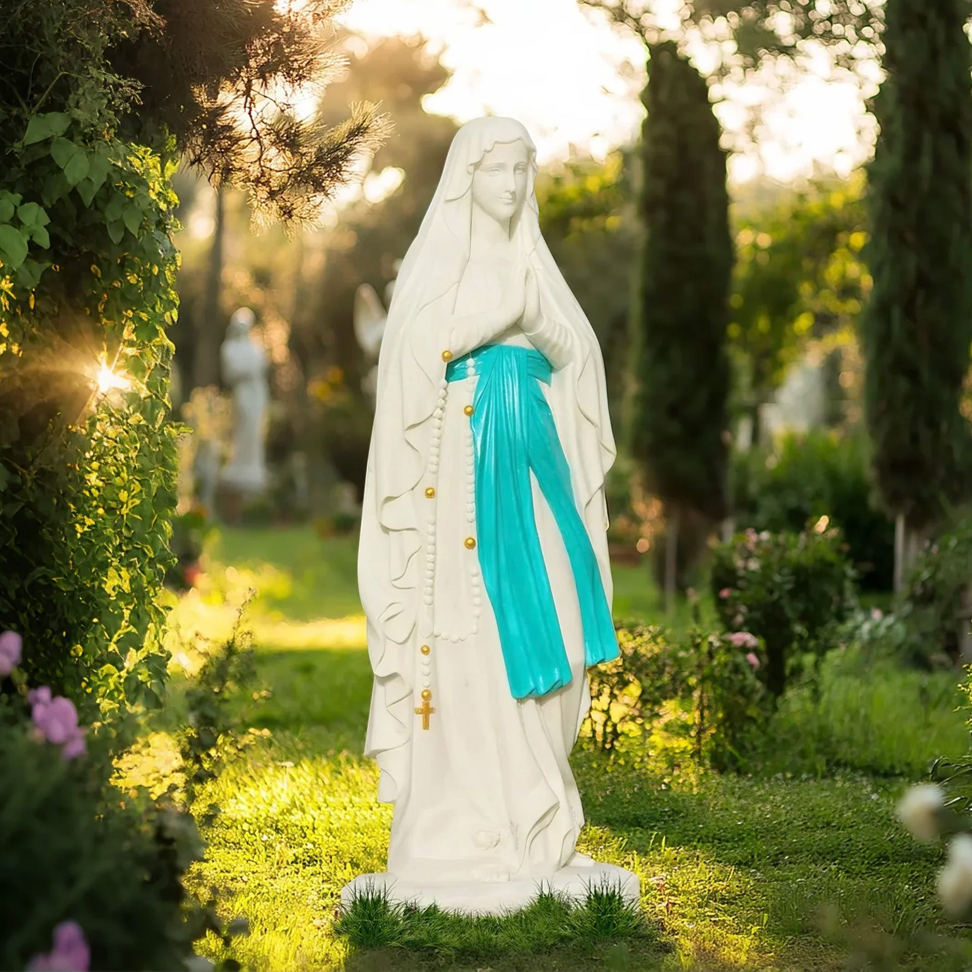 Our Lady of Lourdes white marble statue with painted blue sash, standing in a peaceful garden surrounded by greenery and sunlight.