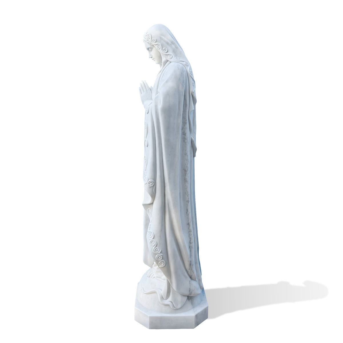 Side profile of a praying Virgin Mary statue in white marble, highlighting her lowered gaze, joined hands and flower-trimmed veil. Deep mantle folds extend toward the feet, with gray veining visible along the side against a white background.