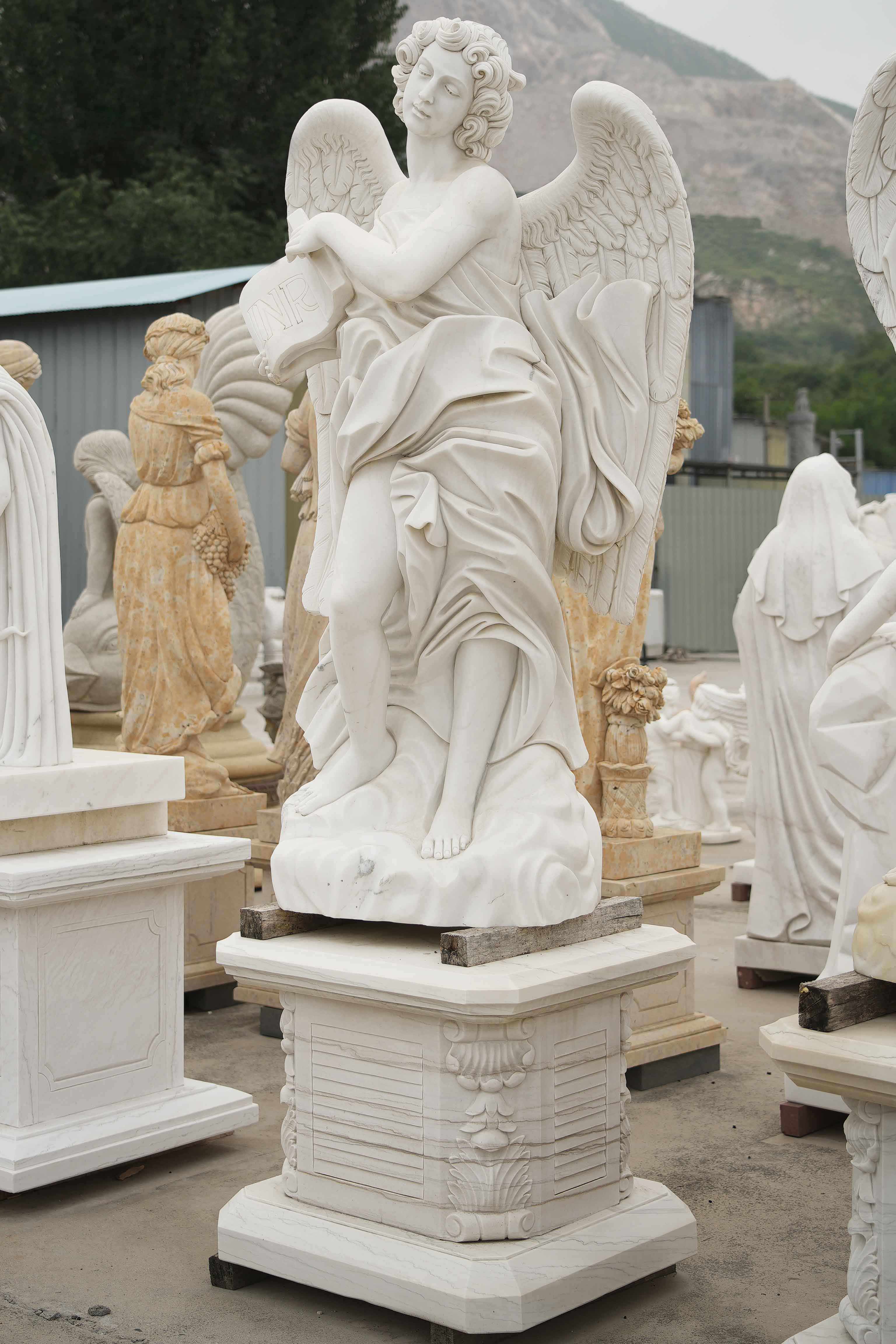 Marble statue of an angel with wings, holding a book inscribed with 'INRI,' in a flowing robe, displayed outdoors among other sculptures.