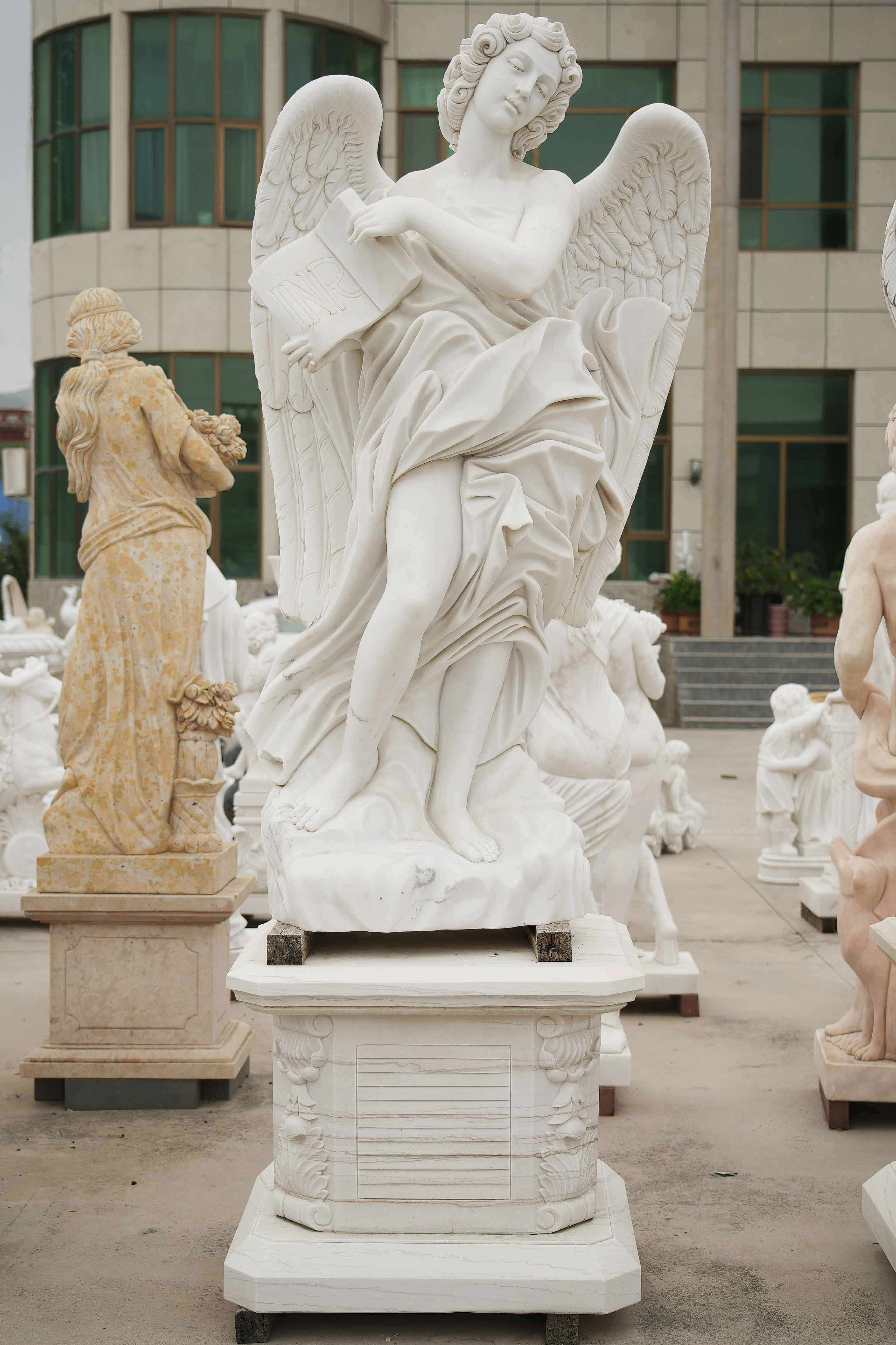 Marble statue of an angel with wings, holding a book inscribed with 'INRI,' in a flowing robe, displayed outdoors among other sculptures.
