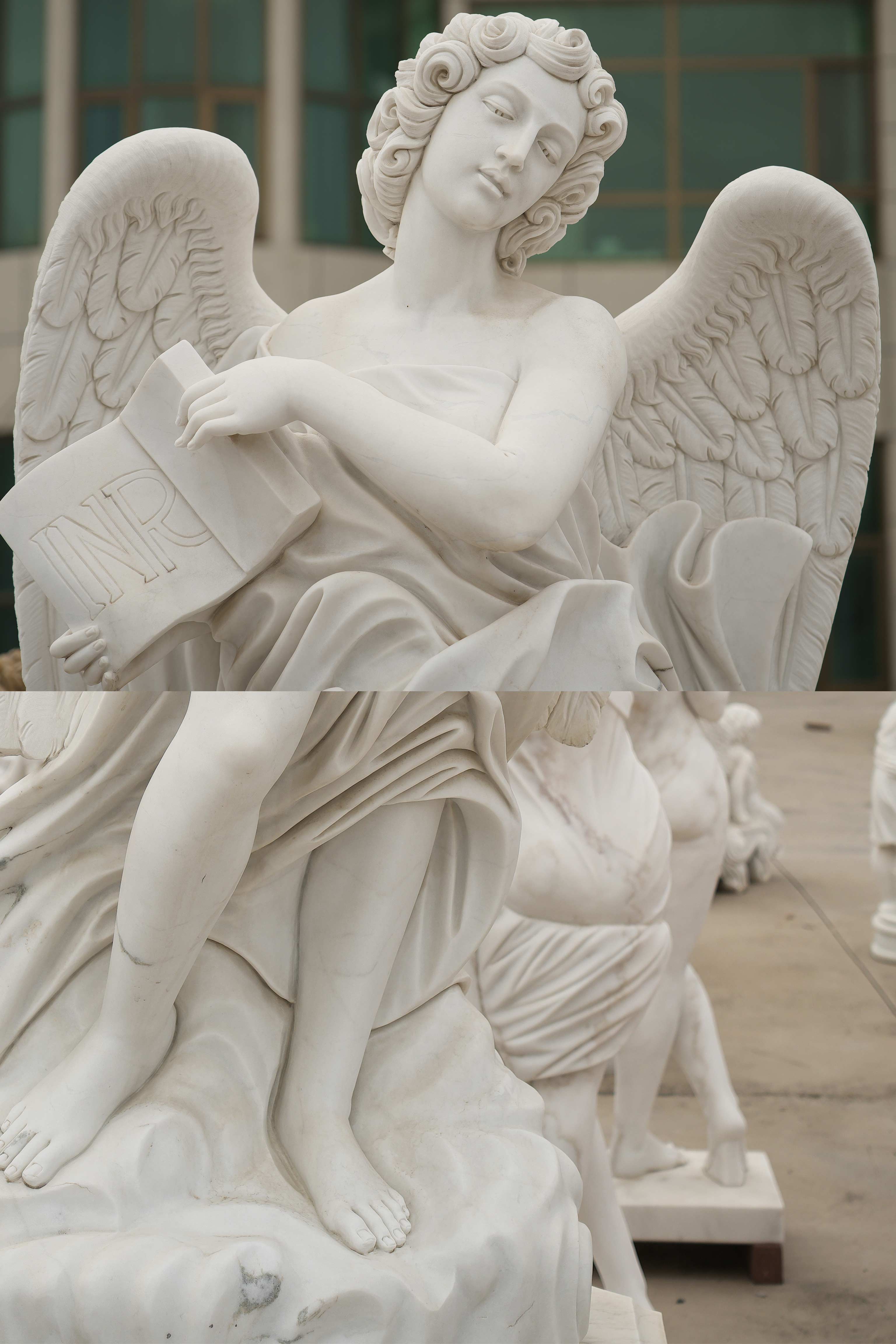 Marble statue of an angel with wings, holding a book inscribed with 'INRI,' in a flowing robe, displayed outdoors among other sculptures.