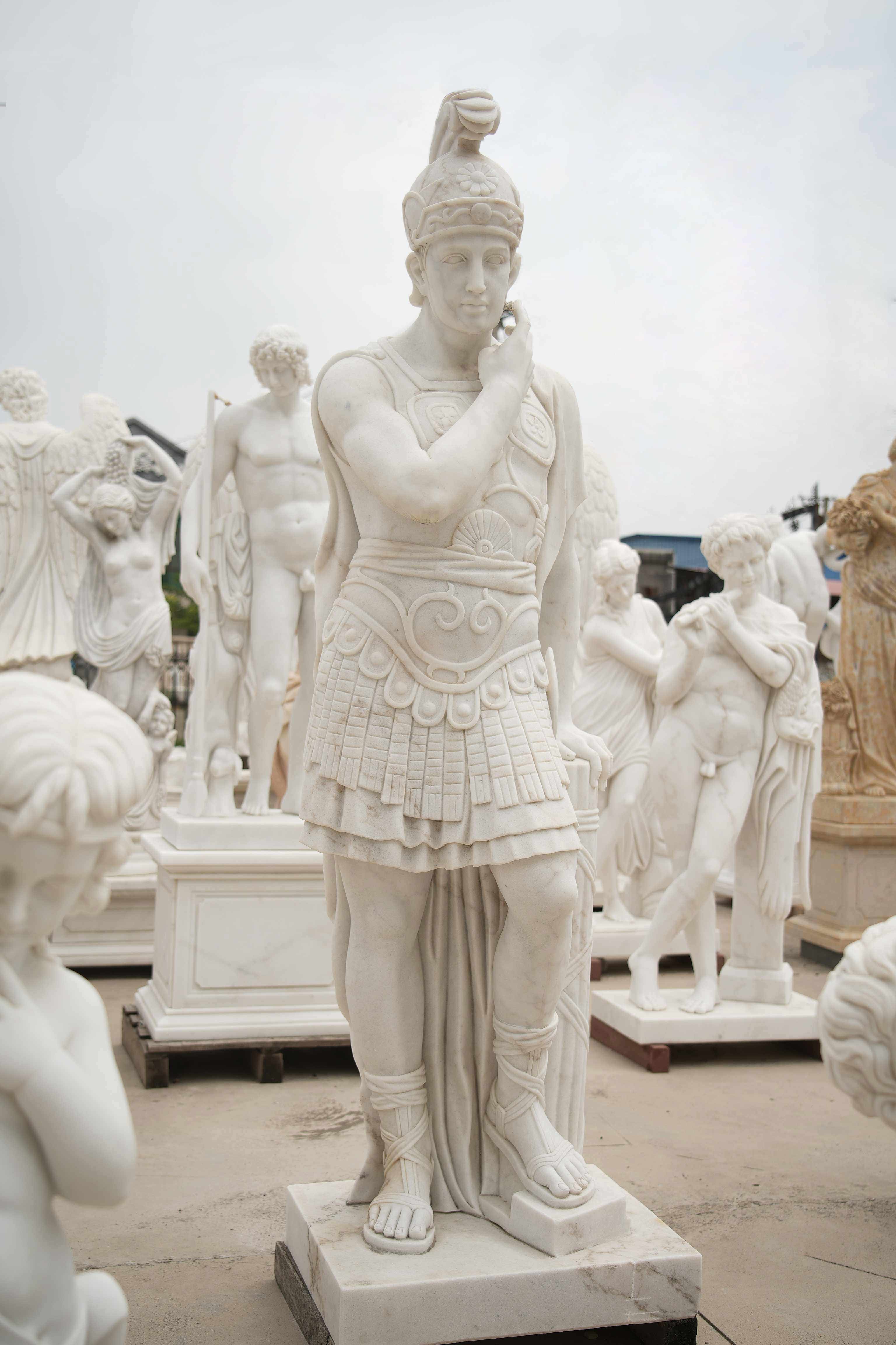 Marble statue of a Roman warrior in a contemplative pose, resting against a pillar, surrounded by other classical sculptures in an outdoor display