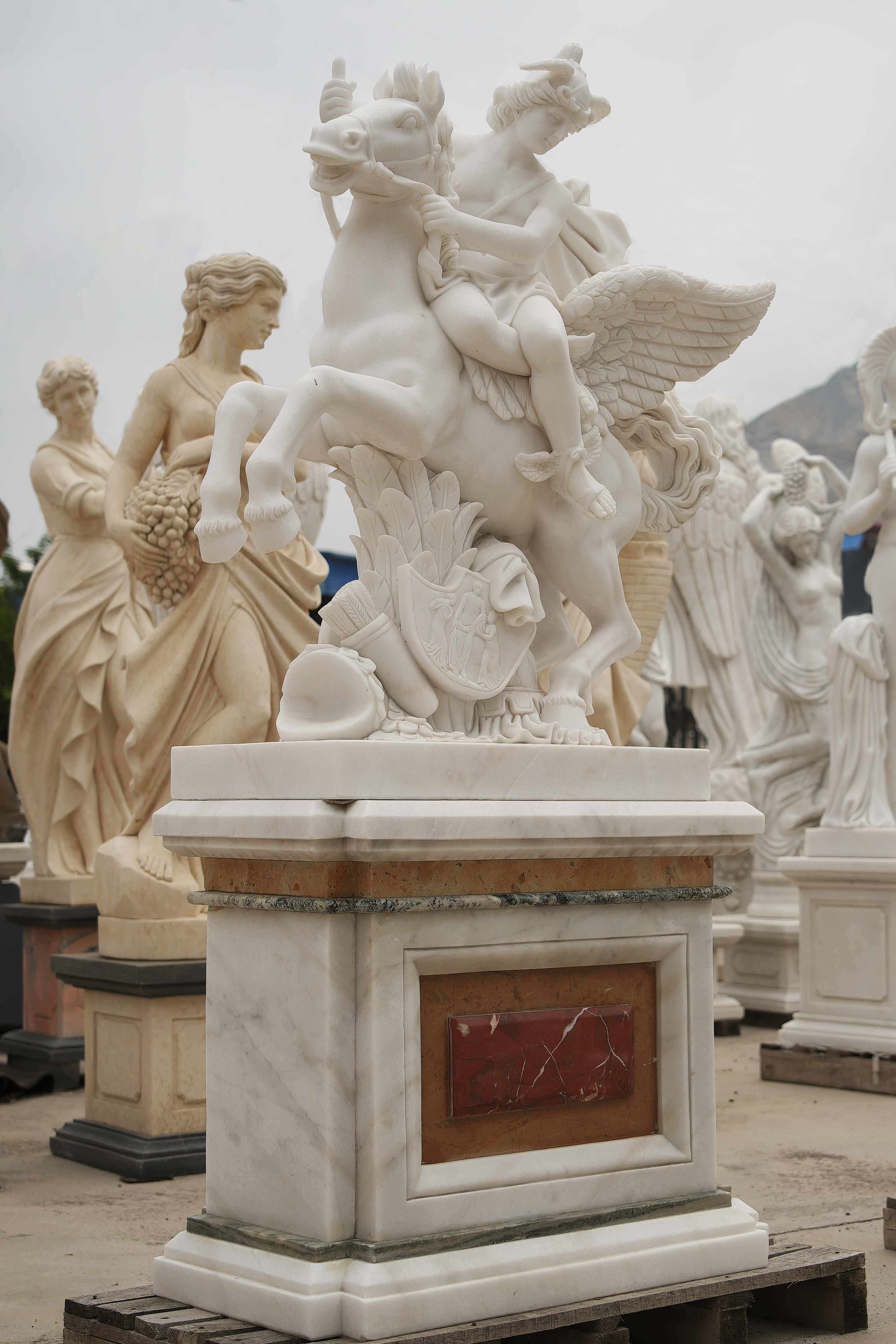 Marble statue of a mythological figure riding a winged horse, detailed with flowing robes, wings, and ornate carvings, surrounded by other classical sculptures.