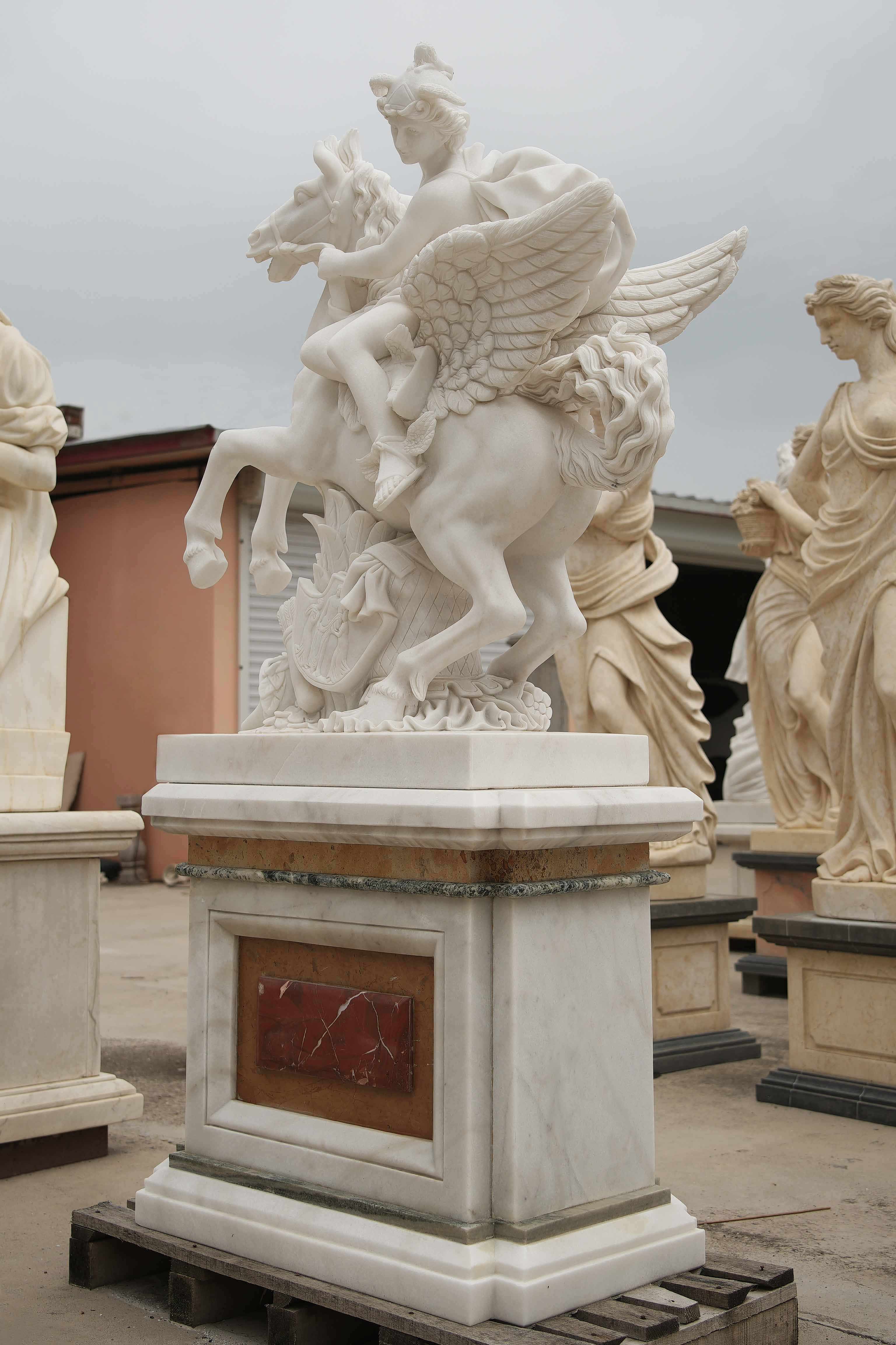 Marble statue of a mythological figure riding a winged horse, detailed with flowing robes, wings, and ornate carvings, surrounded by other classical sculptures.