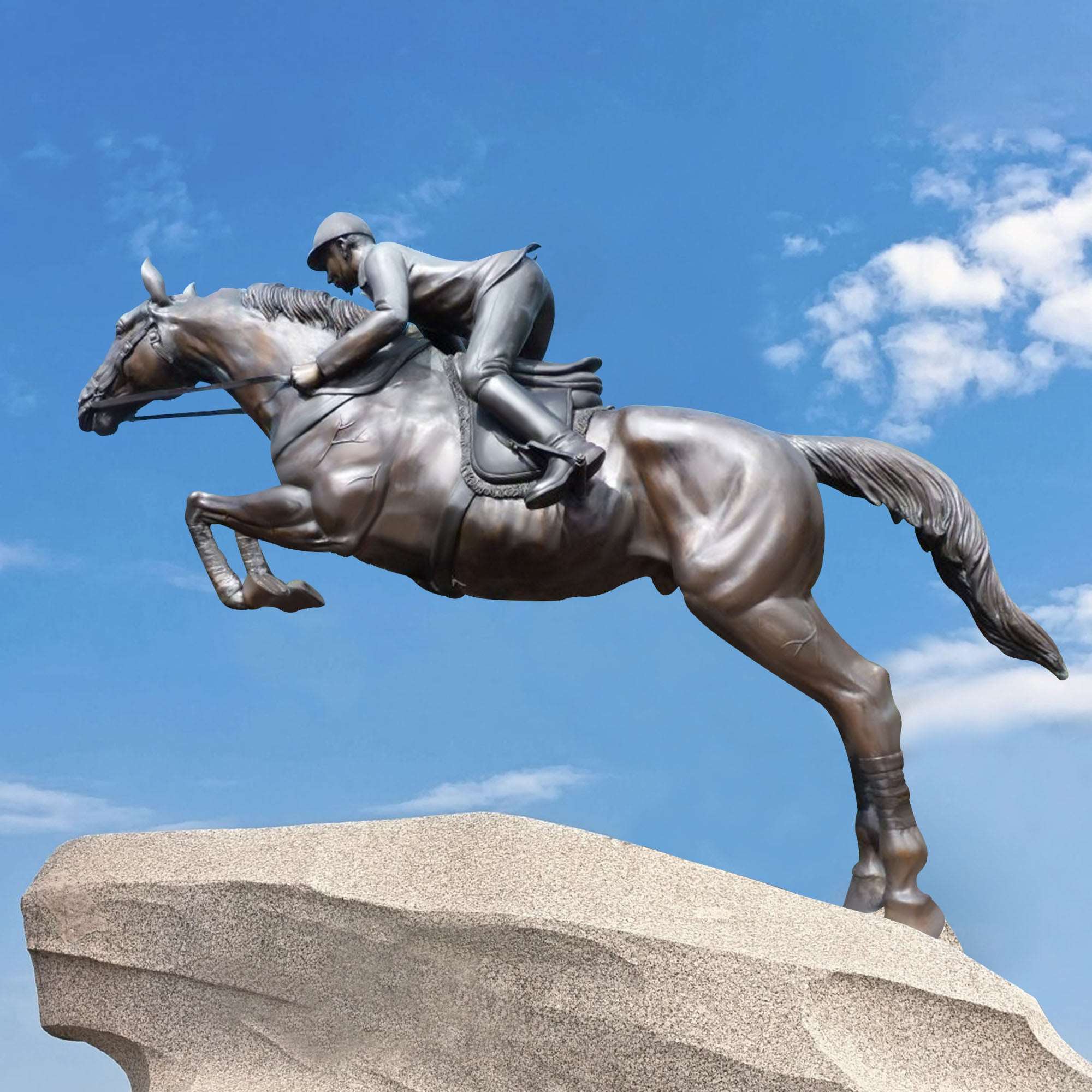 Bronze statue of a horse and rider in mid-jump, mounted on a rugged stone against a clear blue sky