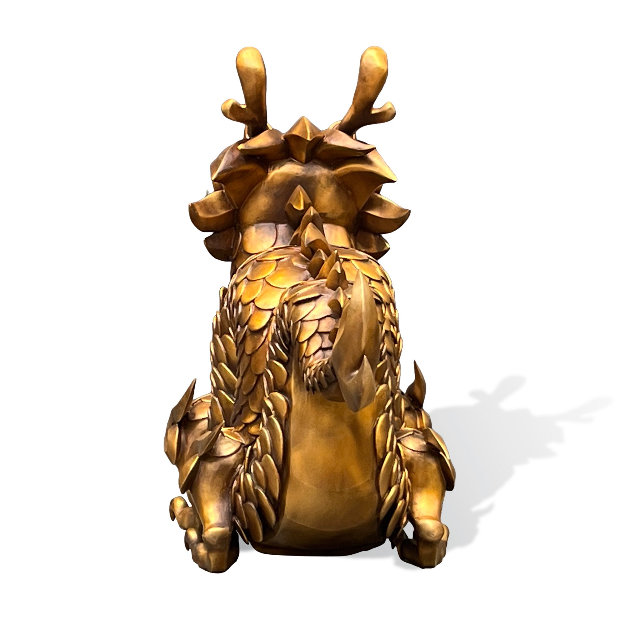 Rear view of a golden bronze dragon sculpture with layered back scales, raised horns, spiked details, curled tail, and strong seated posture on a white background.