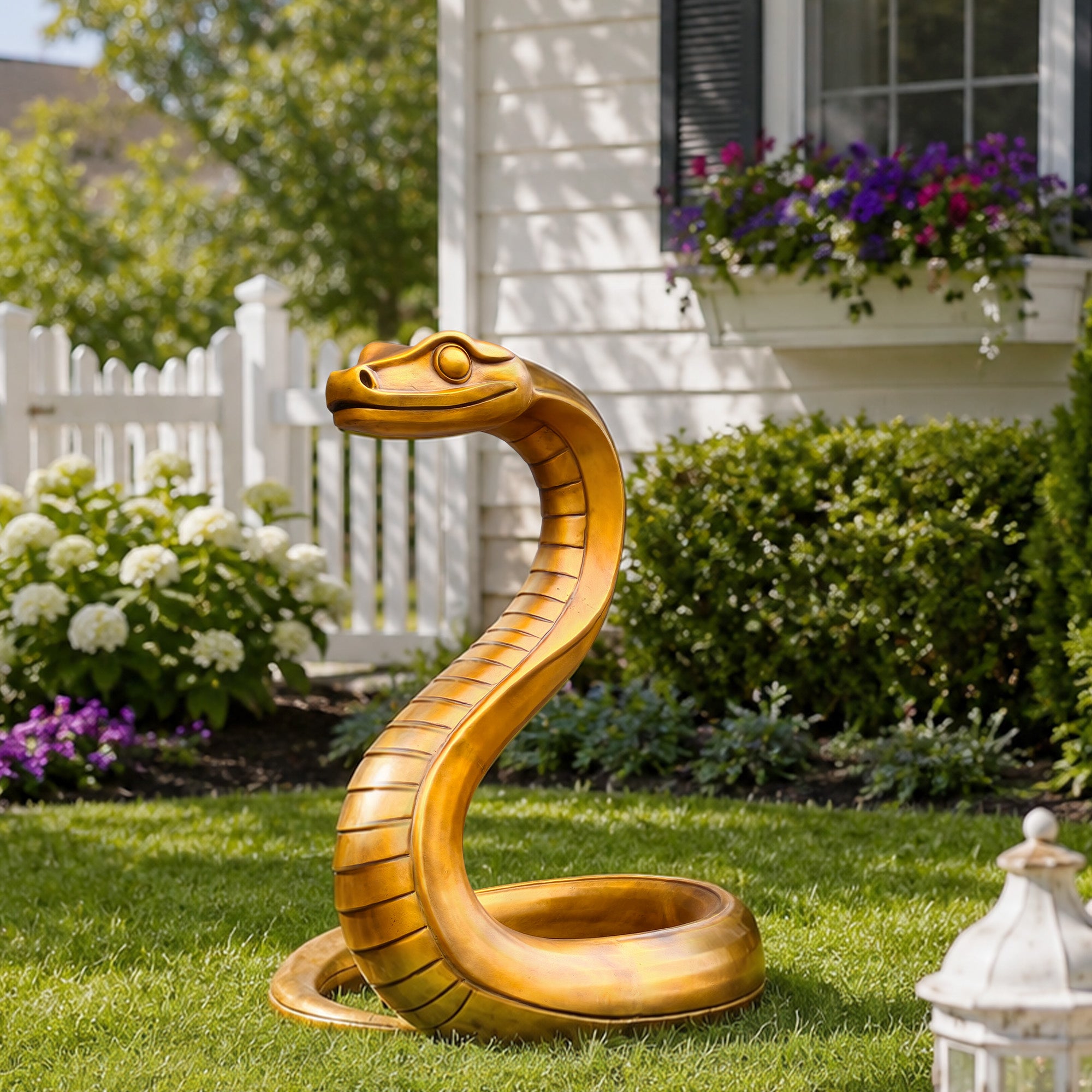 Golden bronze zodiac snake sculpture displayed on a garden lawn with raised cobra-like head, curved body, coiled base, expressive eyes, and landscaped flowers in the background.