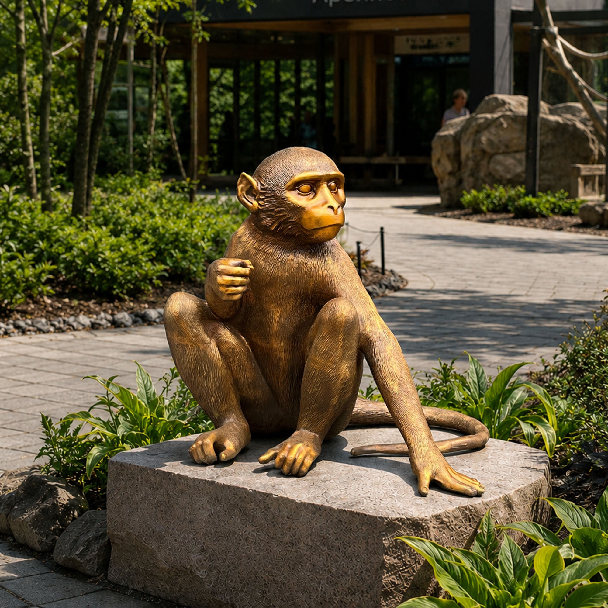 Golden bronze zodiac monkey statue displayed on a stone pedestal in a landscaped garden with seated pose, extended arm, curled tail, textured fur, and outdoor walkway background.