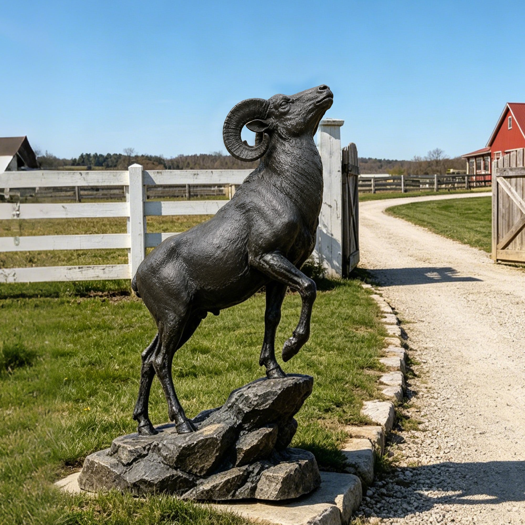 Bronze rearing bighorn ram garden statue displayed on a rock base in a farm landscape, ideal for ranch entrances, estates, parks, and outdoor décor.