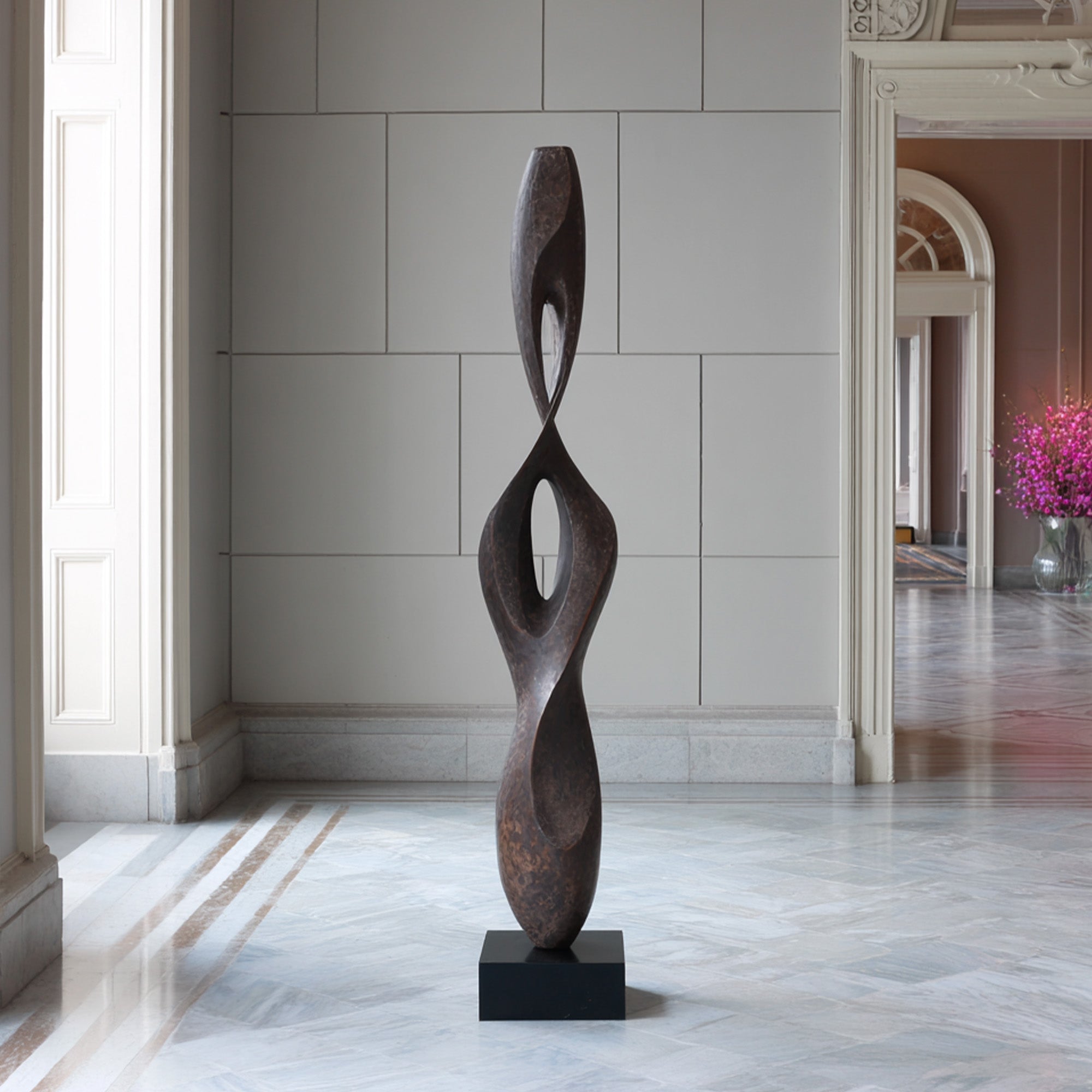 Tall abstract bronze sculpture displayed in a luxury interior, featuring twisting open form, dark textured patina, and black base for modern art decor