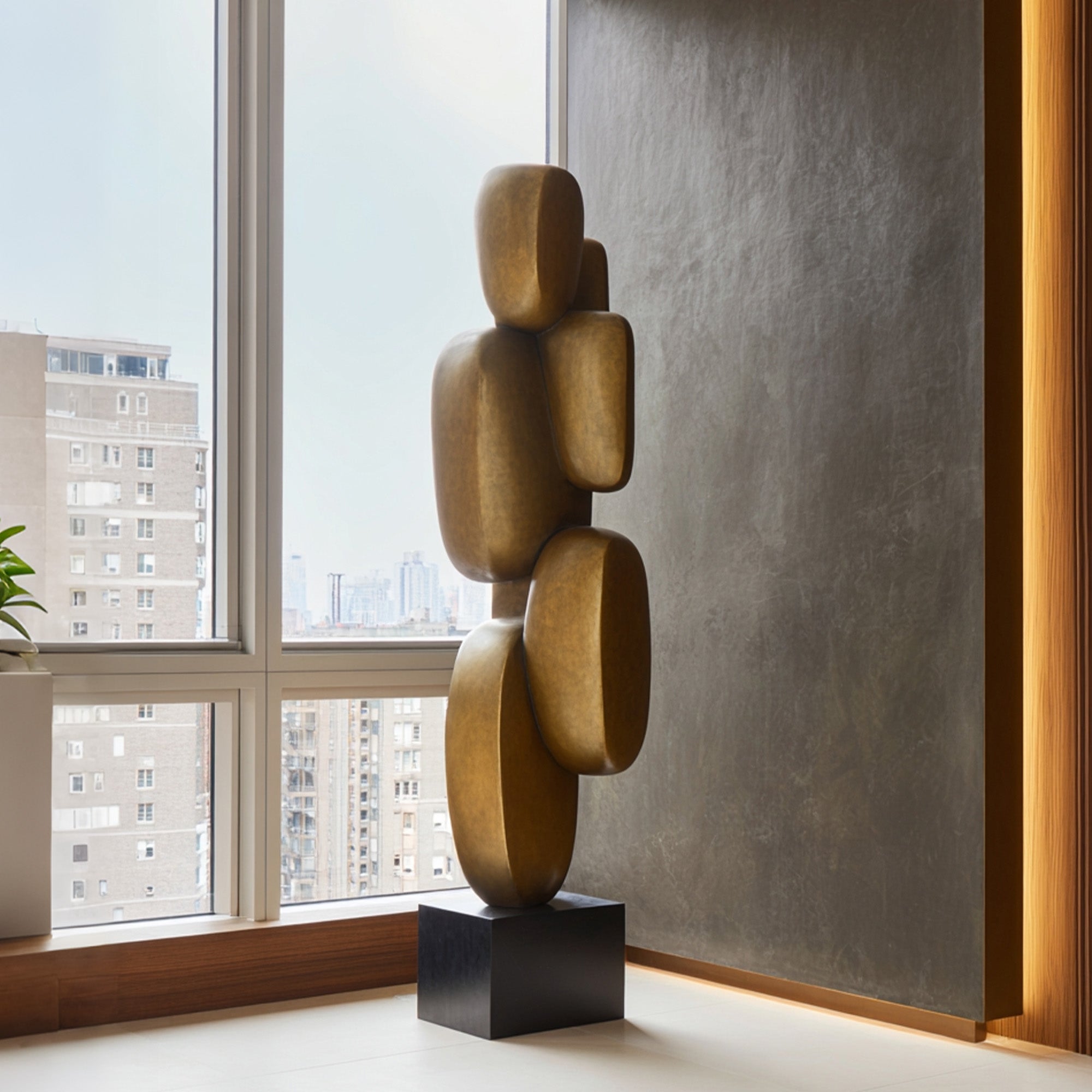 Modern abstract stacked bronze sculpture displayed in a luxury interior by a window, featuring rounded vertical forms, warm bronze finish, and black pedestal base