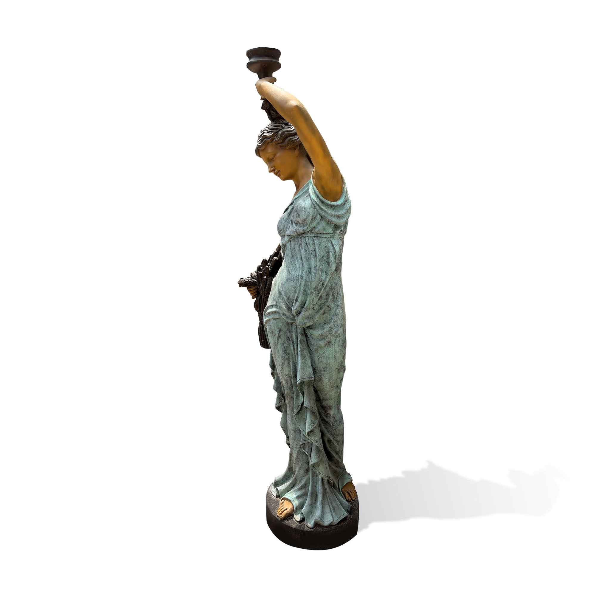 Side view of a classical bronze female figure sculpture with a verdigris patina finish, flowing draped gown, raised vessel, harvest sheaf, and round base.