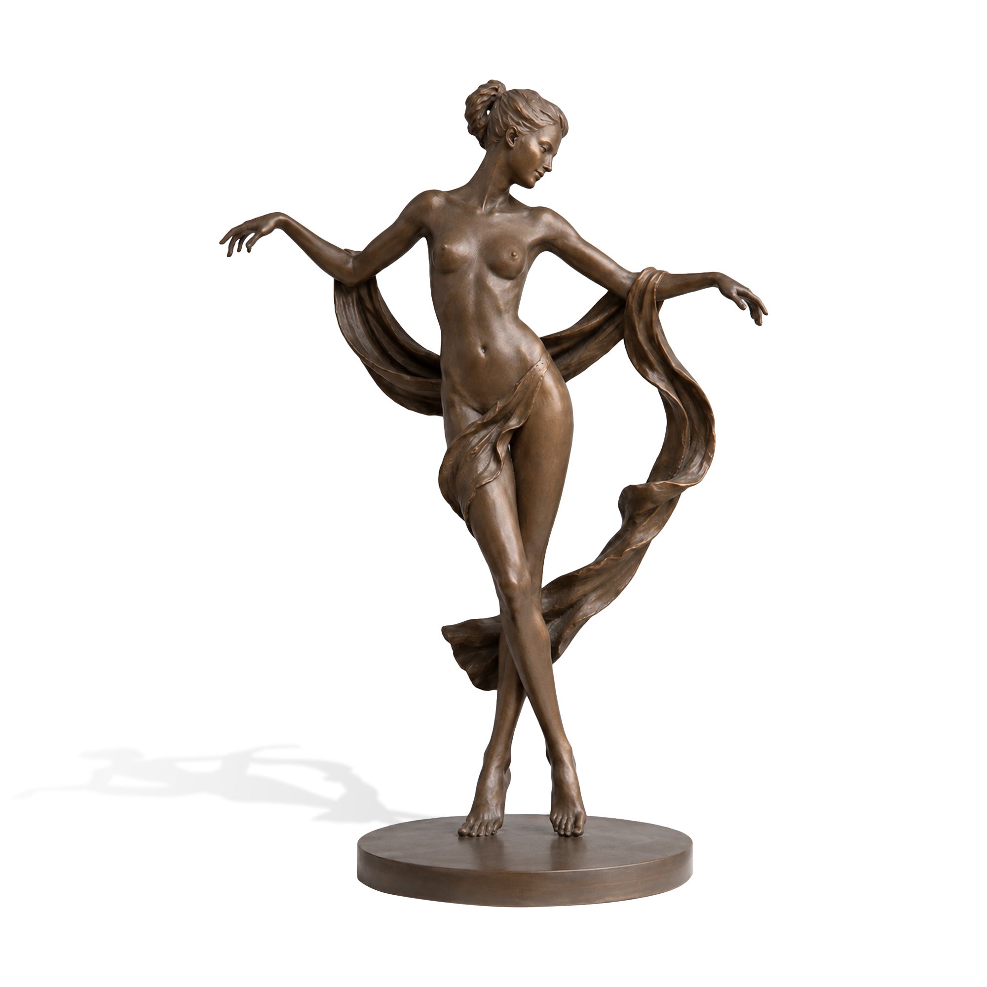 Bronze female dancer sculpture on a white background, depicting a graceful nude woman standing with crossed legs and extended arms, surrounded by flowing drapery that creates a sense of movement, set on a simple round base with a warm brown bronze finish.
