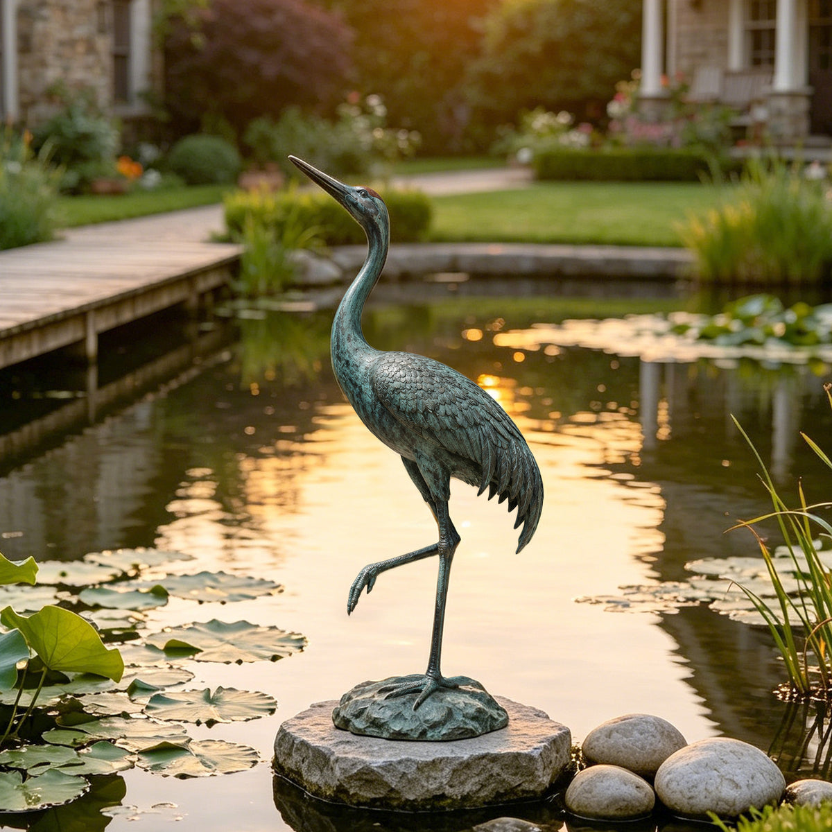 Verdigris bronze crane sculpture standing on one leg beside a garden pond, featuring a long curved neck, raised beak, layered feather details, and a naturalistic rock base surrounded by water lilies and landscaping.