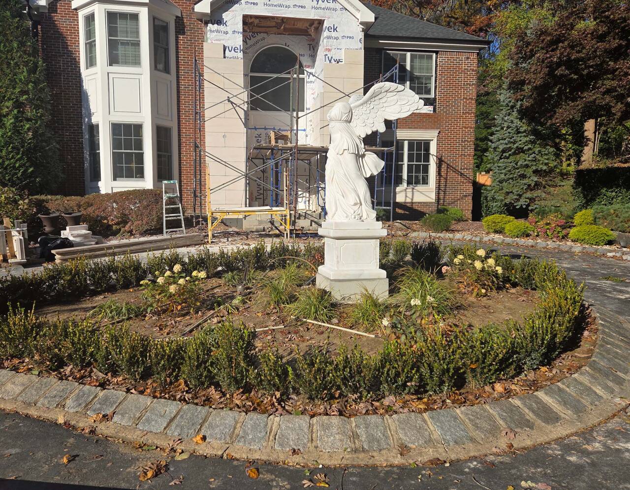 Side view of the marble Winged Victory of Samothrace statue placed in a  garden bed in front of a brick house under construction.
