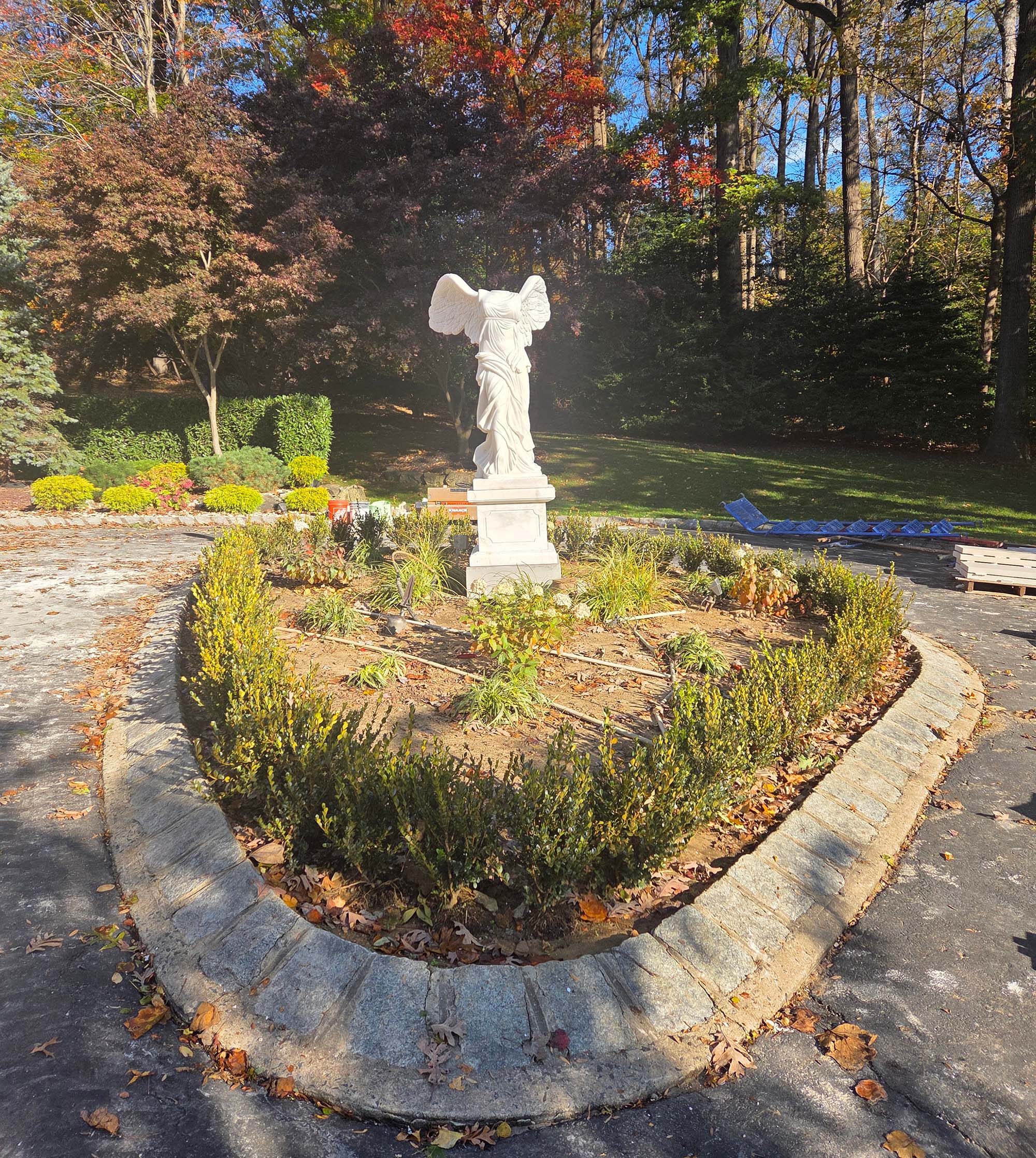 Marble Winged Victory statue installed at the center of a landscaped circular garden bed, surrounded by autumn trees and outdoor greenery.