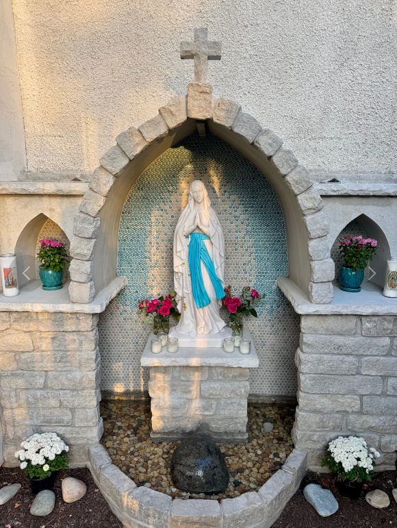 Outdoor Lourdes grotto with a white marble statue of the Virgin Mary draped in a blue sash, displayed inside a stone arch niche with flowers, candles, and a decorative mosaic backdrop.