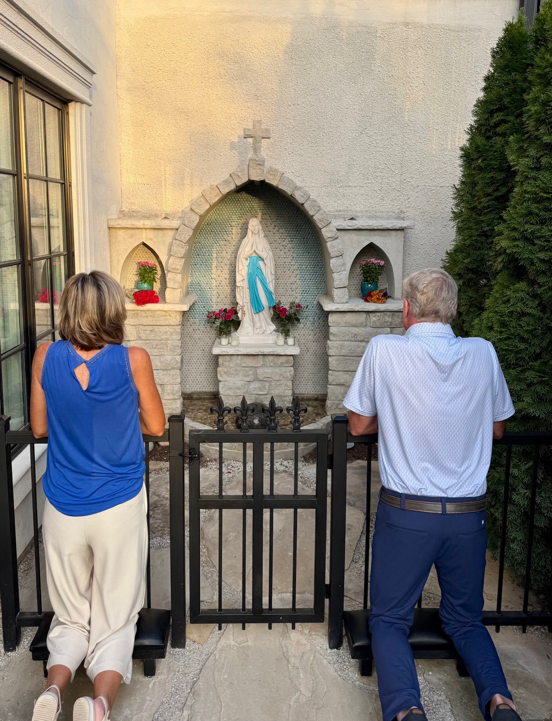 Two people kneeling in prayer in front of an outdoor Lourdes grotto featuring a statue of the Virgin Mary, surrounded by flowers and candles in a peaceful devotional setting.