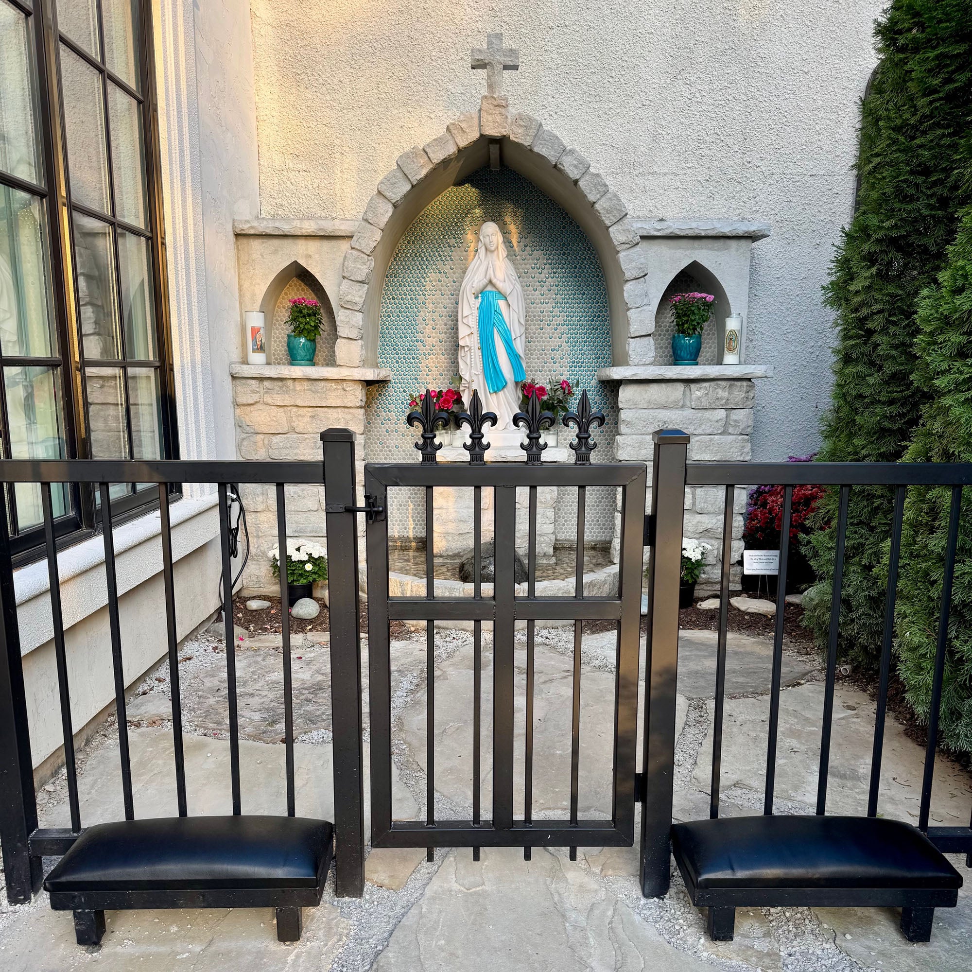 Our lady of Lourdes outdoor Marian grotto with a Virgin Mary statue behind a black metal prayer rail and kneelers, framed by stone arches, flowers, and a serene garden setting.