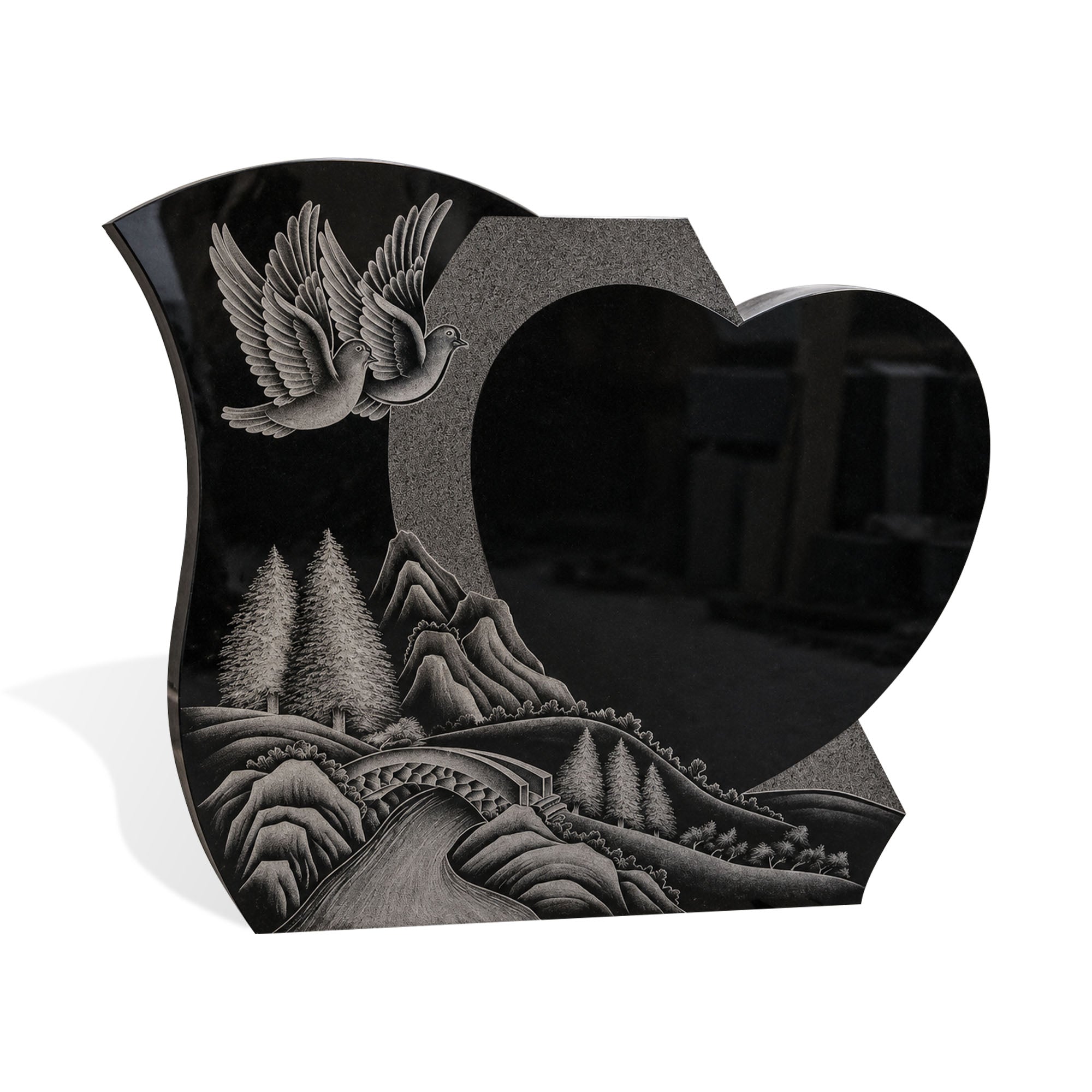 Black granite heart-shaped memorial headstone with engraved doves, mountain scenery, arched bridge, pine trees, polished black surface, and white background