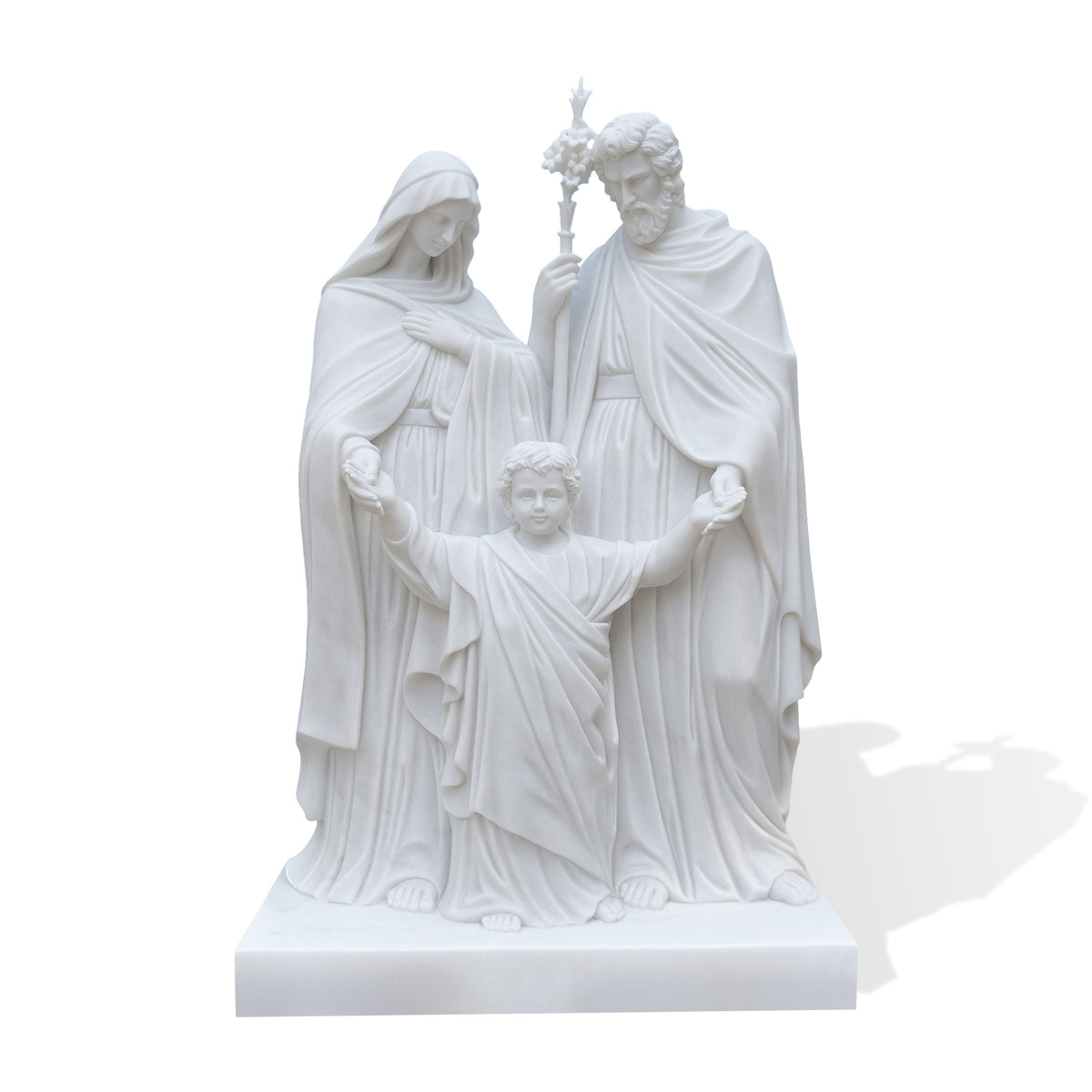 White marble Holy Family statue displayed outside a church, featuring the Virgin Mary, Saint Joseph with a flowering staff, and the child Jesus standing between them with raised arms.