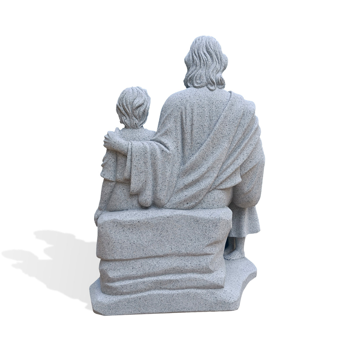 Rear view of a gray granite Jesus with children statue, showing carved wavy hair, flowing robe folds and an arm resting around a child’s shoulders. The figures sit on a rock-like bench above an irregular stone base against a white background.