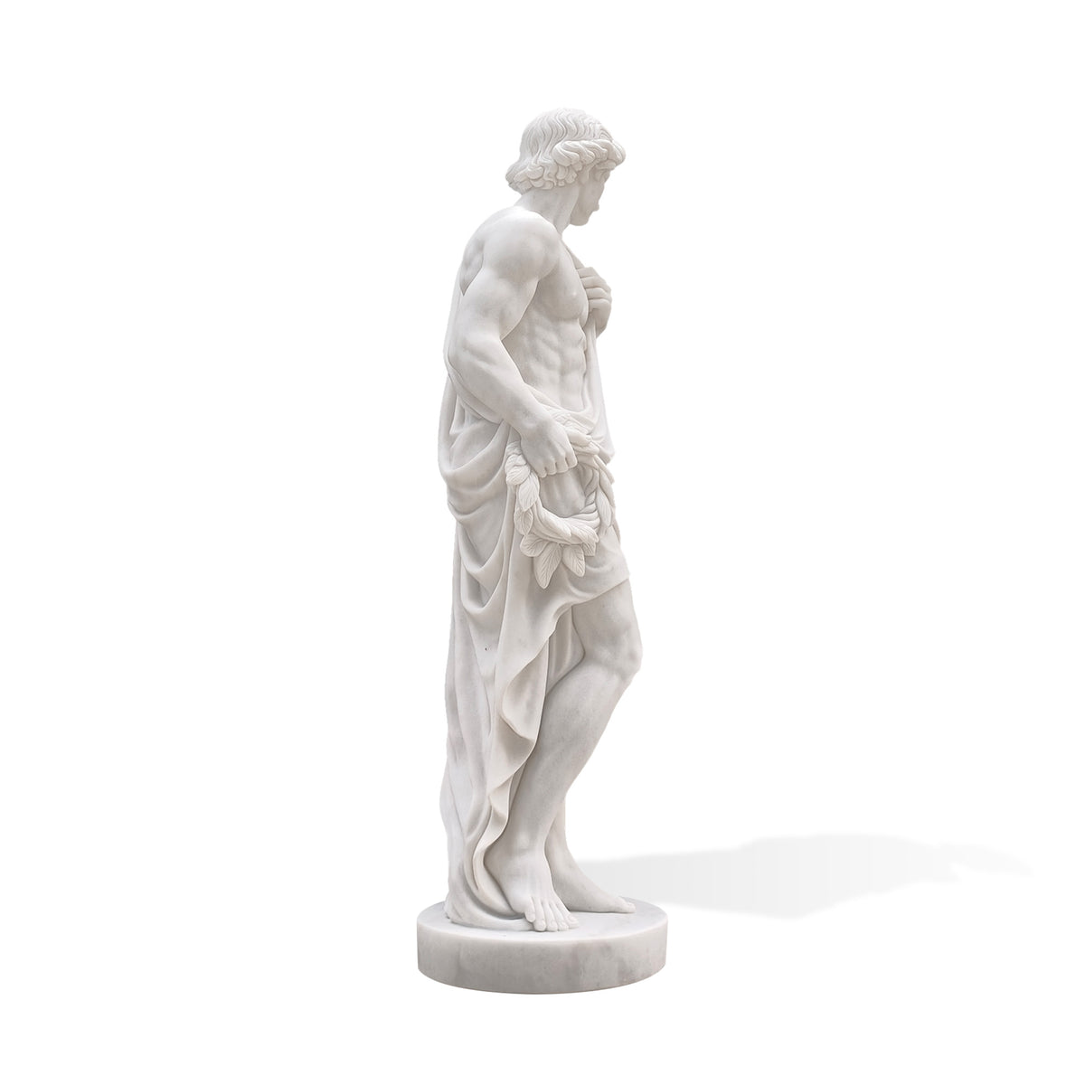 Side view of a white marble male statue highlighting the carved arm and torso, a leafy laurel wreath held at the hip and cascading drapery behind the legs. The figure’s lowered gaze and shifted stance are visible above a circular marble base.