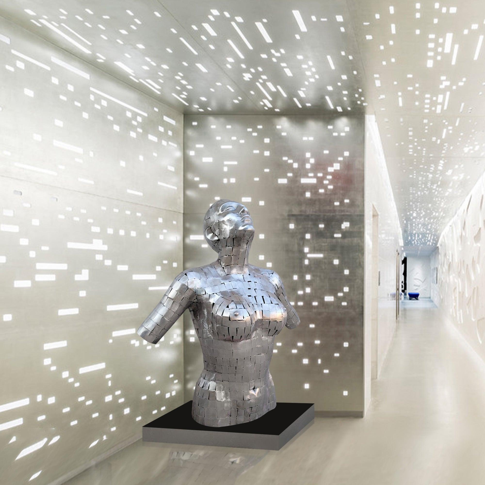 A polished stainless steel sculpture of a female bust with an abstract mosaic design, mounted on a black base, situated in a modern, illuminated hallway with geometric light patterns on the walls and ceiling.