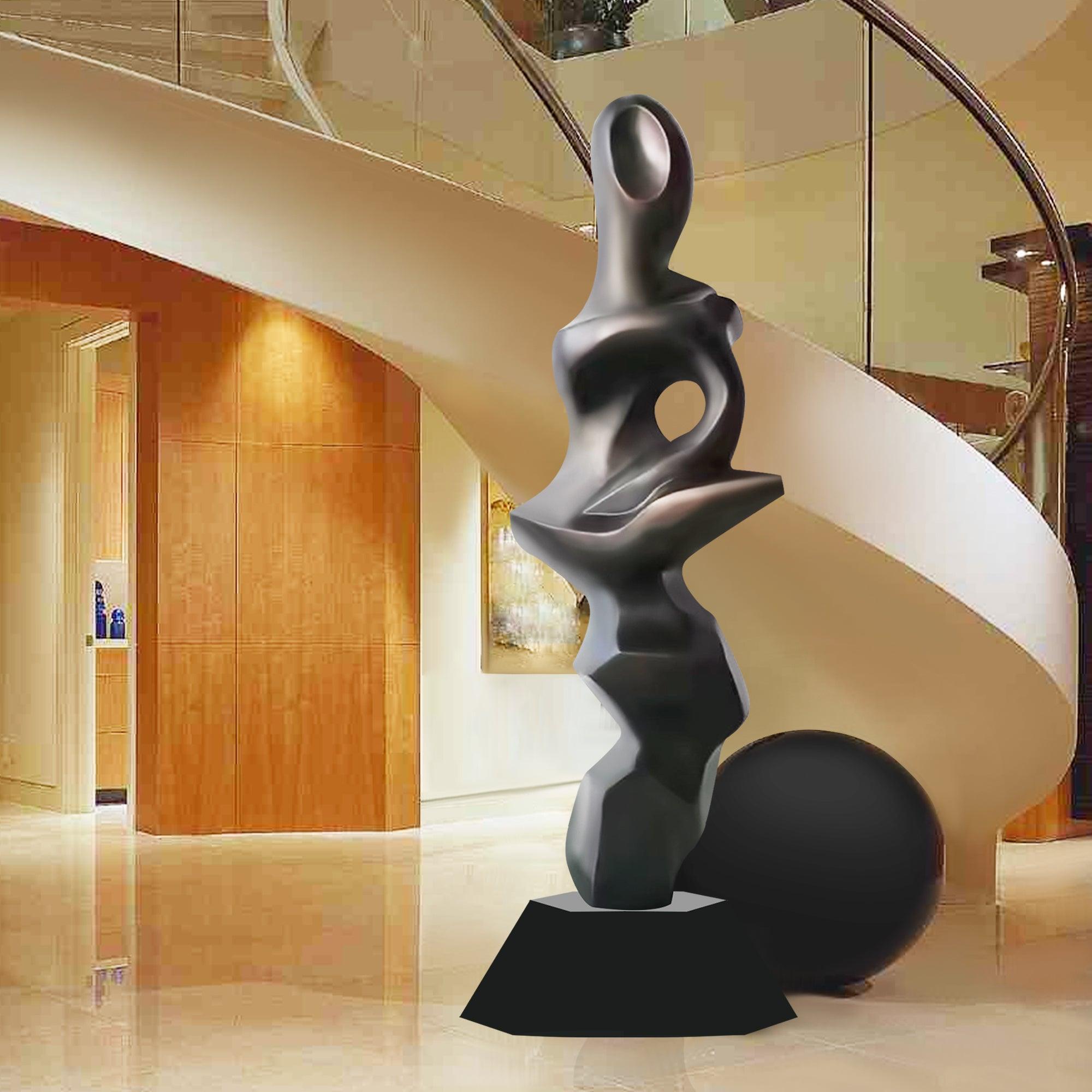 Abstract stainless steel floor sculpture with fluid, organic shapes and a matte finish, standing on a sturdy black base in a modern interior with a spiral staircase and wooden accents.