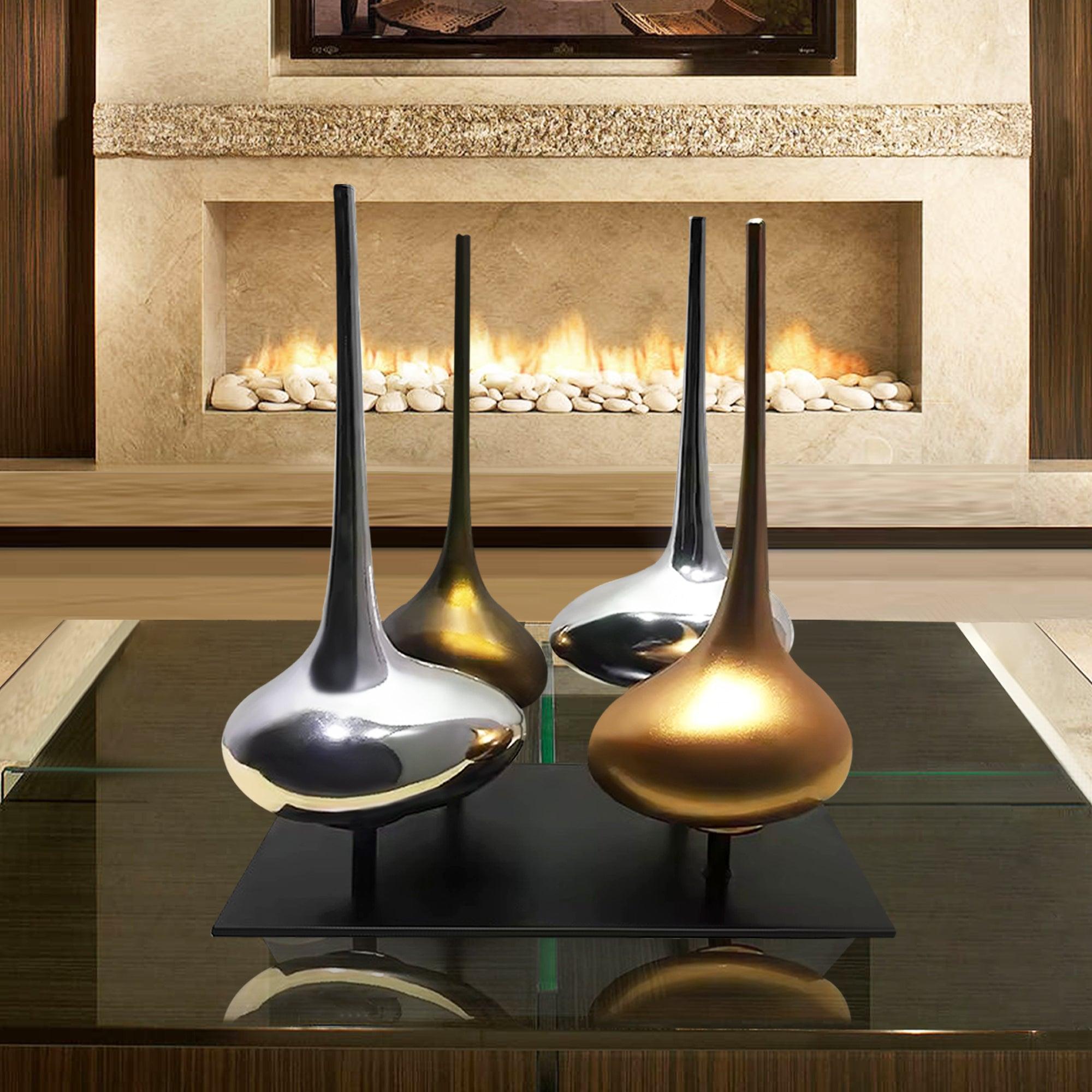 Modern metal flasks sculptures in silver and gold finishes, mounted on a black base, displayed on a glass table in front of a beige stone fireplace with white pebbles and a glowing fire.