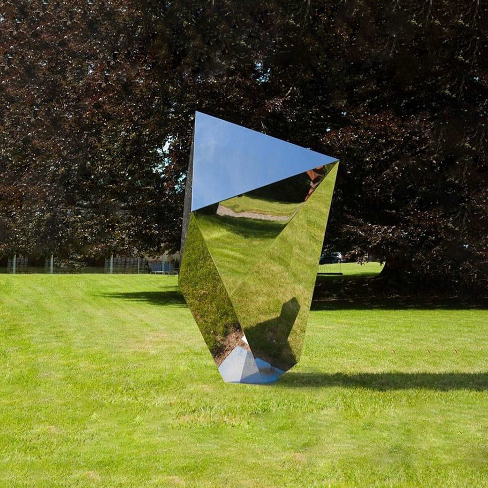 Large polished stainless steel geometric block sculpture with a mirror surface, displayed on a green lawn, reflecting the surrounding trees and sky