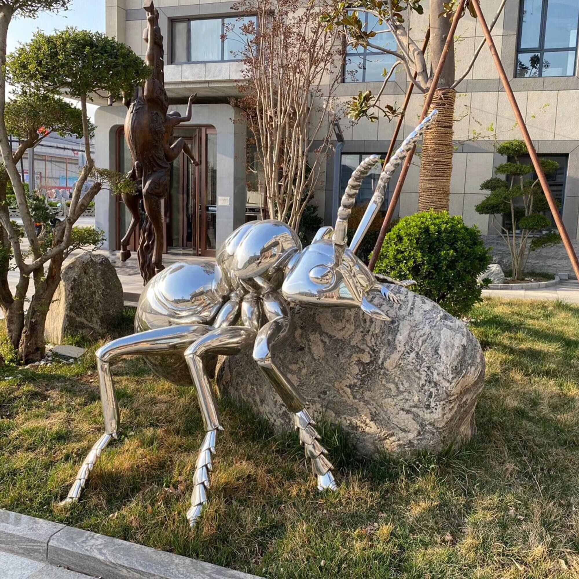 Stainless steel ant animal sculpture placed outdoors next to a rock, reflecting the surrounding environment and showcasing intricate, polished details