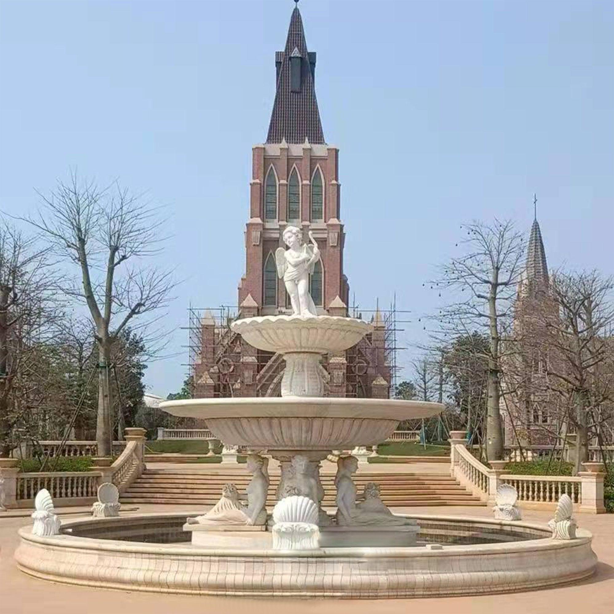 2 tiers white statue fountatin in public garden with angle standing in top and two women below