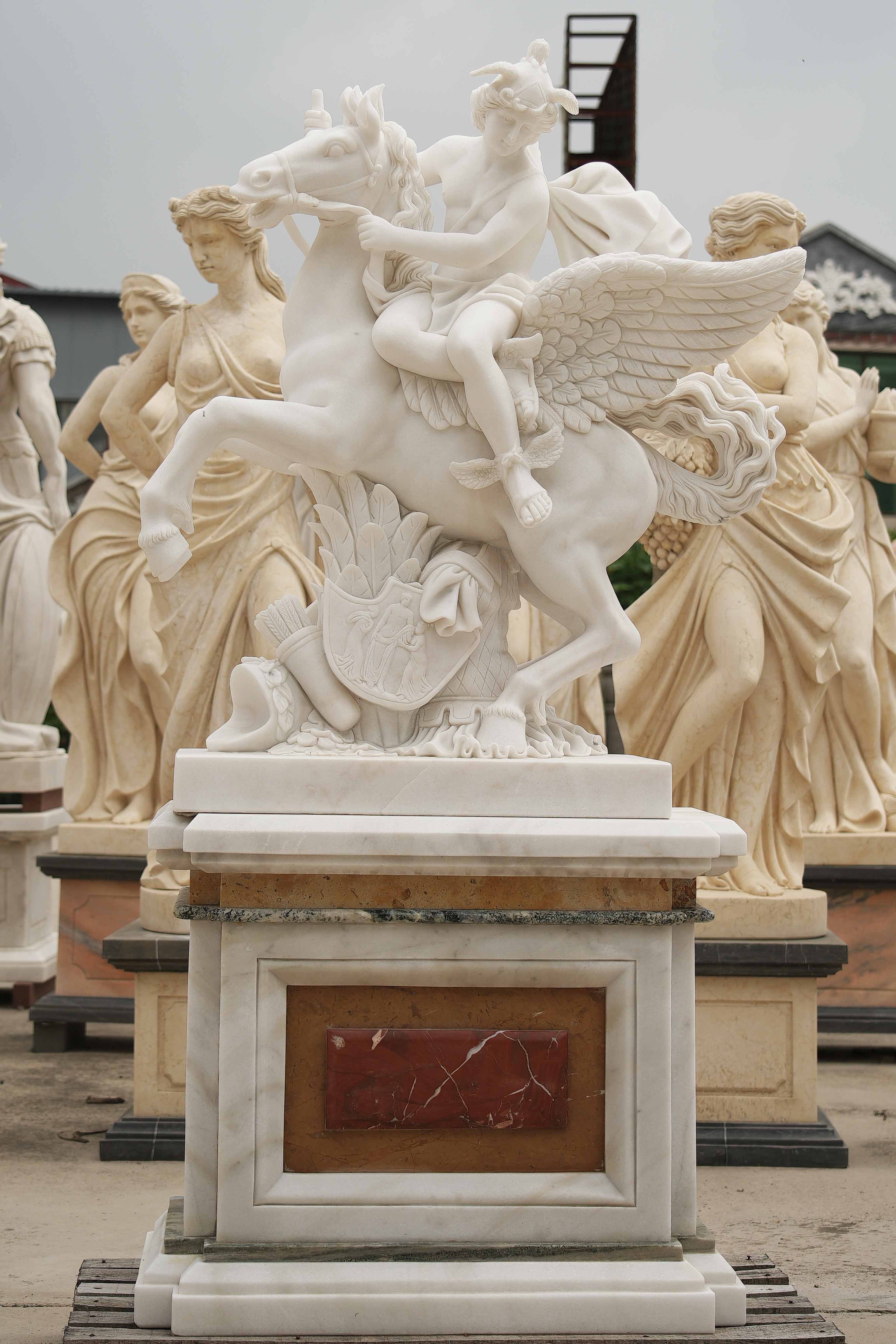 Marble statue of a mythological figure riding a winged horse, detailed with flowing robes, wings, and ornate carvings, surrounded by other classical sculptures.